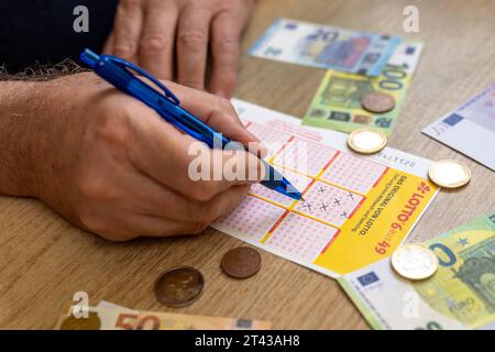 Augusta, Baviera, Germania - 27 ottobre 2023: Un uomo compila un biglietto della lotteria della lotteria 6 su 49 con una penna a sfera. Accanto a lui ci sono diverse banconote e monete in euro. Immagine simbolo lotto milionaire and profit *** Ein Mann füllt einen Lottoschein von der Lotterie 6 aus 49 mit einem Kugelschreiber aus. Neben IHM liegen Mehrere Euro Geldscheine und Geldmünzen. Symbolbild lotto Millionär und Gewinn Foto Stock