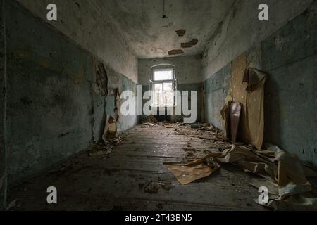 L'interno della camera abbandonata con pareti dipinte che si staccavano danneggiava la spazzatura dei mobili di legno sul pavimento sporco con soffitto sgretolato che mostrava aste di ferro Foto Stock
