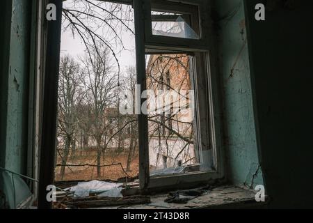Vecchia vetrina con telaio in metallo rotto con pareti sporche e sporche, con reti di ragno nere distrutte e affacciate sul verde edificio in mattoni di alberi d'autunno, sotto il cielo blu senza nuvole di giorno Foto Stock