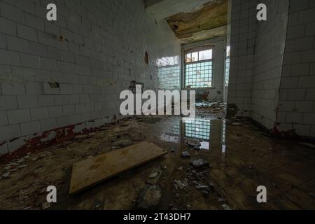 Interno di mattonelle in ceramica rotte abbandonate con mobili rotti di macerie sparse e acqua riflettente sul pavimento alla luce del giorno attraverso finestre con recinzione Foto Stock