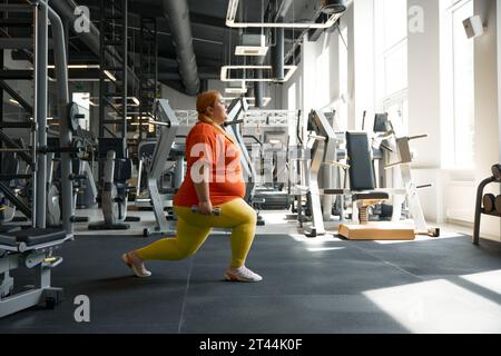 Donna obesa sicura che fa affondi in avanti con manubri in palestra Foto Stock