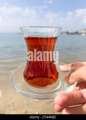 Tè, vista laterale, le donne tengono a mano un bicchiere di tè turco o alle erbe sulla splendida spiaggia sabbiosa e il cielo blu in una giornata soleggiata con spazio fotocopie. Turco tradizionale Foto Stock