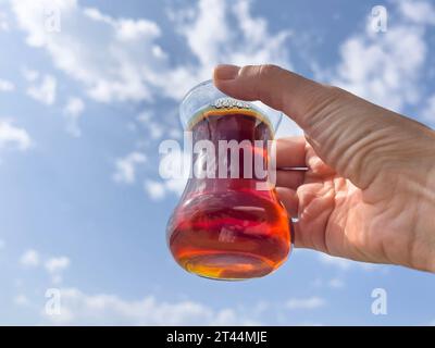 Tè, mano femminile ad angolo basso tiene un bicchiere di tè turco o alle erbe isolato su un bel cielo blu con soffici nuvole bianche in una giornata di sole con spazio per le copie. Foto Stock