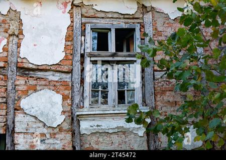 Finestra verde d'epoca con vetro frantumato su una casa maledetta, che rivela mattoni e travi rossi. Un rustico giorno autunnale in campagna. Foto Stock