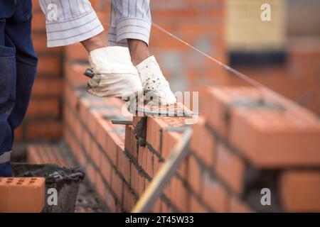 Primo piano delle mani di un costruttore di guanti pone un muro di mattoni su una corda con cazzuola Foto Stock