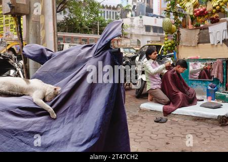 Street Life India: Un parrucchiere in una strada a Mumbai, India, che lavora su un cliente proprio sul passaggio pedonale, mentre un gatto dorme su una motocicletta nelle vicinanze Foto Stock