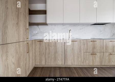 Angolo di una cucina moderna con mobili in legno chiaro, maniglie e rubinetti in metallo dorato, ripiano in quarzo imitato installato in un grande appartamento con un Foto Stock