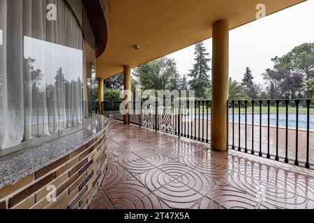 Portico coperto di una casa unifamiliare con pavimento in terrazzo rosso, ringhiere metalliche e piscina all'aperto nel giardino pieno di erba e alberi di varie varietà Foto Stock