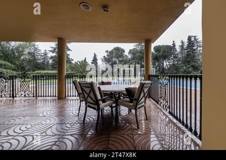 Portico coperto di una casa unifamiliare con pavimento in terrazzo rosso, ringhiere in metallo, tavolo circolare con sedie in metallo con cuscini e piscina all'aperto in sala Foto Stock