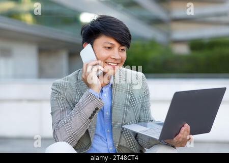 Uomo d'affari asiatico che parla al telefono navigando sul sito Web su un computer portatile all'aperto Foto Stock