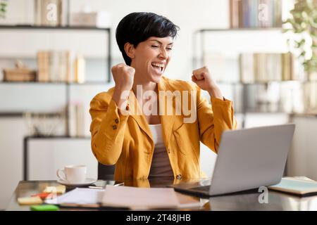 Emotiva bruna signora al laptop che scuote le puzze al tavolo da lavoro Foto Stock