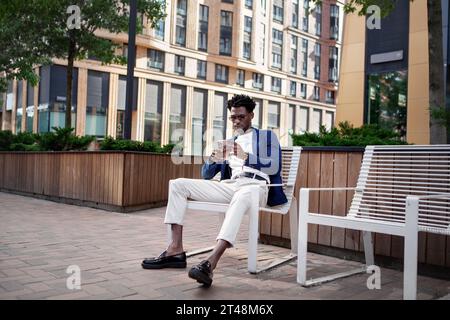Uomo nero a figura intera seduto sulla panchina in un ambiente urbano e guarda il telefono, la digitazione o la navigazione. Ragazzo vestito con stile in abito con giacca e pelo bianco Foto Stock
