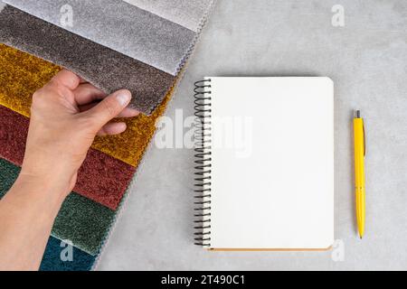 Campioni di tessuti colorati, blocco note e penna su tavolo bianco. La mano tocca i campioni di tessuto, impostati in diversi colori per la selezione. Varietà di tappezzeria m Foto Stock