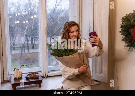 Elegante donna con maglione bianco scatta selfie sullo smartphone, tiene in mano il bouquet natalizio in carta artigianale vicino alla finestra. Giovane ragazza millenaria che ha f Foto Stock