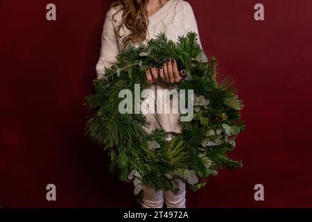 Ritratto ravvicinato giovane donna senza volto con maglione bianco a maglia tiene in mano la corona di Natale verde su sfondo scuro e isolato. Posto per Foto Stock