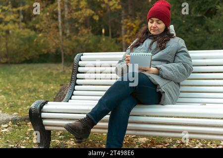 Donna di mezza età che si gode l'atmosfera tranquilla del parco, seduto su una panchina, creando meravigliosi schizzi nel suo notebook Foto Stock