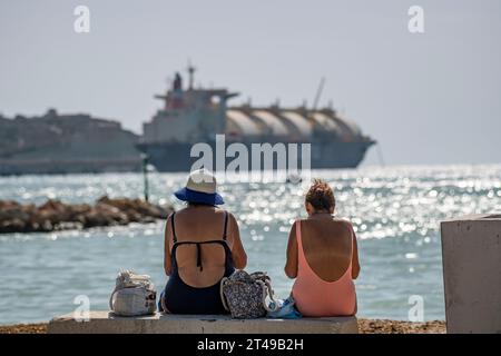 Due signore in costume da bagno si godono la spiaggia con l'Armada LNG Mediterrana in lontananza, un'unità galleggiante di stoccaggio e rigassificazione di gas a Malta. Foto Stock