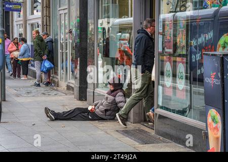 Un senzatetto siede di fronte al negozio in strada in attesa di donazioni da parte dei passanti. Dublino, Irlanda. Foto Stock