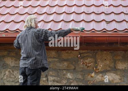 Un uomo maturo rimuove foglie e detriti dalla fossa della sua casa. Pulizia di uno scarico della pioggia. Foto Stock