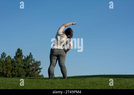 la donna di mezza età in sovrappeso fa esercizi nel parco in piedi su erba, sfondo blu cielo, vista posteriore. la donna grassa inclina il corpo da un lato all'altro. Stile di vita sano Foto Stock