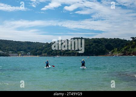 Due paddle boarder si dirigono lontano dalla spiaggia di Sunny Cove Beach, East Portlemouth, in una giornata estiva dal cielo blu, verso North e South Sands, Salcombe. Foto Stock