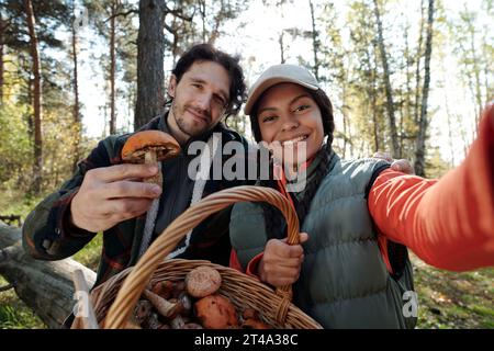 Un paio di giovani raccoglitori di funghi sorridenti con cesto pieno di vari funghi che guardano la fotocamera mentre scattano selfie nella foresta autunnale Foto Stock