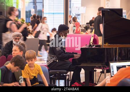 Montreal, Quebec, Canada - 07 04 2022: Un musicista adolescente che indossa una felpa nera Unbreakable con cappuccio che suona il pianoforte a coda nero in mezzo a un affollato e affollato outbound Foto Stock