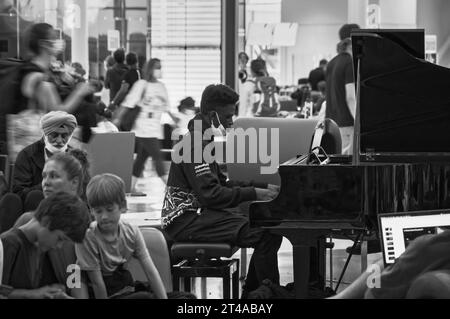 Montreal, Quebec, Canada - 07 04 2022: Un musicista adolescente che indossa una felpa nera Unbreakable con cappuccio che suona il pianoforte a coda nero in mezzo a un affollato e affollato outbound Foto Stock