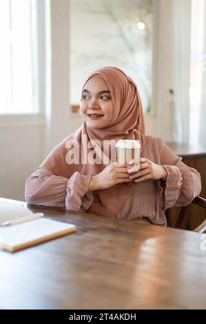 Un ritratto di una bella e rilassata donna asiatica-musulmana che indossa un hijab sta guardando lontano dalla macchina fotografica, sorridendo e sognando ad occhi aperti la sua felicità Foto Stock