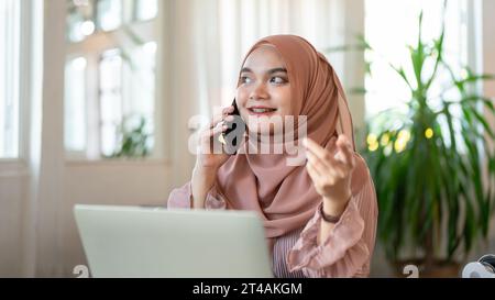 Una giovane donna asiatica-musulmana attraente e allegra chiama qualcuno o parla al telefono con qualcuno mentre lavora in remoto in un bar. P Foto Stock