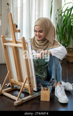 Una giovane artista asiatica-musulmana bella e felice è seduta sul pavimento e si diverte a creare le sue opere d'arte su una tela, dipingendo nel suo studio d'arte. Foto Stock