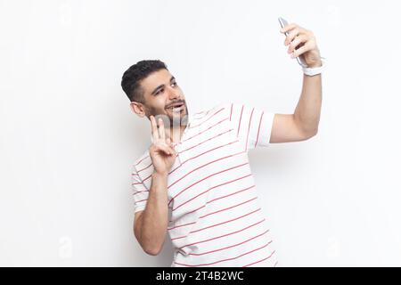 Ritratto di un uomo gioioso, giocoso e barbuto che indossa una t-shirt a righe in piedi che fa selfie, mostrando un V sign ai suoi seguaci. Riprese in studio in interni isolate su sfondo grigio. Foto Stock