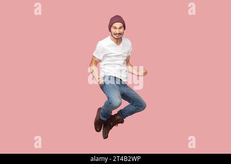 Ritratto di un uomo barbuto estremamente felice e gioioso in T-shirt bianca e cappello di fagiolo che salta con pugni stringuti, gioisce e celebra la vittoria. Riprese in studio per interni isolate su sfondo rosa. Foto Stock