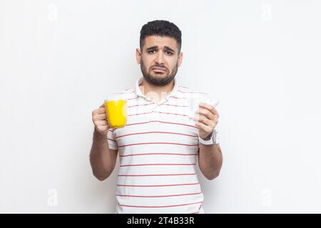 Ritratto di un uomo barbuto triste infelice e malsano che indossa una t-shirt a righe in piedi con una tazza gialla, che guarda la macchina fotografica, si sente male, il volto si sbaglia. Riprese in studio in interni isolate su sfondo grigio. Foto Stock