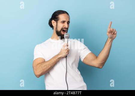 Ritratto di un uomo buffo con la barba che indossa una T-shirt bianca che canta ad alta voce una canzone che tiene il microfono in mano, divertendosi a riposare nel karaoke e a cantare. Riprese in studio in interni isolate su sfondo blu Foto Stock