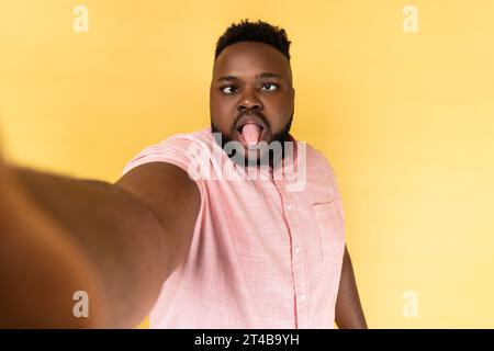 Ritratto di un divertente uomo barbuto infantile che indossa una camicia rosa che scatta foto dal punto di vista, guardando la macchina fotografica con gli occhi incrociati e la lingua fuori. Riprese in studio in interni isolate su sfondo giallo. Foto Stock