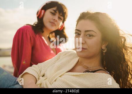 Due donne, una indossata in rosso brillante con le cuffie e l'altra con un maglione in avorio, condividono un momento di tranquillità in riva al mare, riflettendo il calore del Foto Stock