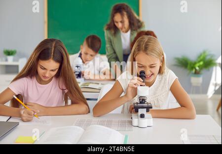 Studentessa che lavora in classe con il microscopio mentre la sua compagna di classe prende appunti durante una lezione Foto Stock