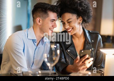 Un giovane uomo e una donna, seduti in un ristorante chic, condividono un momento di gioia guardando uno smartphone. I loro sorrisi e la loro vicinanza suggeriscono un profondo connectio Foto Stock
