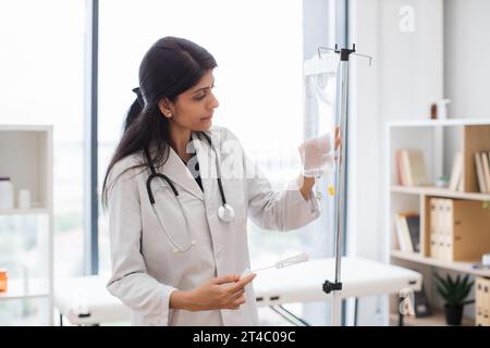 Medico femminile adulta in camice da laboratorio bianco in piedi vicino al contagocce medico e controllo farmaci liquidi. Donna indiana che prepara infusione goccia per intrav Foto Stock