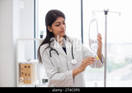 Medico femminile adulta in camice da laboratorio bianco in piedi vicino al contagocce medico e controllo farmaci liquidi. Donna indiana che prepara infusione goccia per intrav Foto Stock