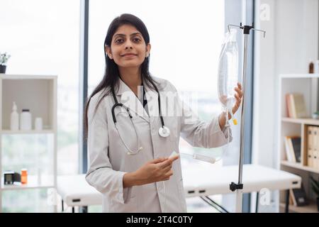 Medico femminile adulta in camice da laboratorio bianco in piedi vicino al contagocce medico e controllo farmaci liquidi. Donna indiana che prepara infusione goccia per intrav Foto Stock