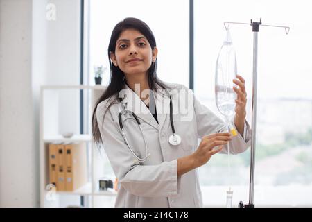 Medico femminile adulta in camice da laboratorio bianco in piedi vicino al contagocce medico e controllo farmaci liquidi. Donna indiana che prepara infusione goccia per intrav Foto Stock