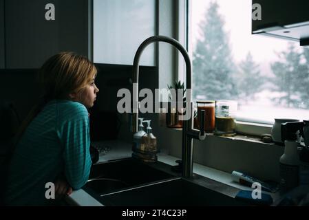 La ragazza preadolescente si appoggia sul lavandino della cucina e guarda fuori dalla finestra di sno Foto Stock