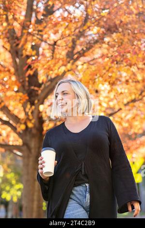 Donna alla moda che indossa il nero, tiene una tazza di caffè e cammina in autunno Foto Stock