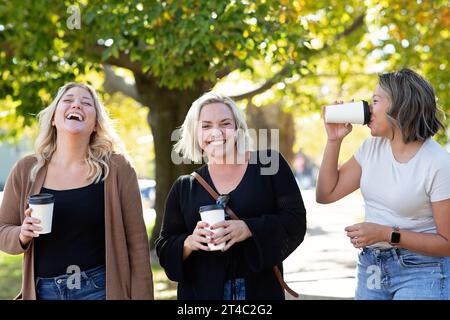 Tre donne che tengono tazze di caffè che camminano, ridono Foto Stock