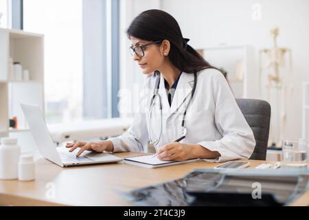 Vista laterale del medico femminile indiano utilizzando il computer portatile mentre si siede alla scrivania dell'ufficio all'interno del centro di salute. Pratica generale efficiente e matura Foto Stock