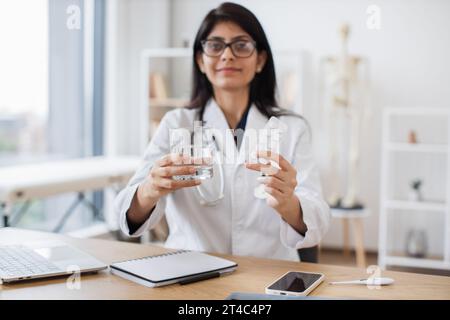 Concentratevi su un piatto di pillole che tiene da una donna indiana seduta alla scrivania con un portatile sullo sfondo dell'ufficio. Operatore medico professionale che dà m Foto Stock