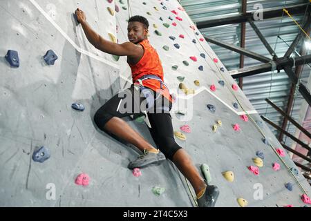 forte uomo afroamericano in camicia arancione che sale su un muro con corda di sicurezza e guarda in basso Foto Stock