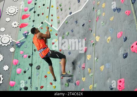 atletico afro-americano con camicia arancione appeso alla sua corda da arrampicata e guardando in alto, parete di roccia Foto Stock